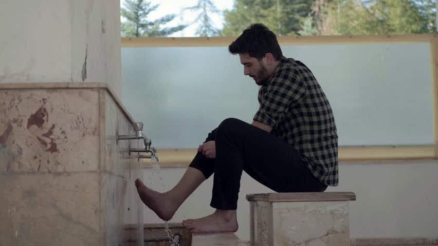 Young Muslim man taking ablution for prayer the mosque, side view teenager washing his feet in the fountain