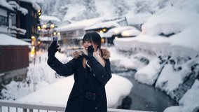 Asian woman using mobile phone taking selfie during travel at Ginzan Onsen area street in Yamagata prefecture, Japan covered in snow. Attractive girl travel local village landmark on winter vacation.