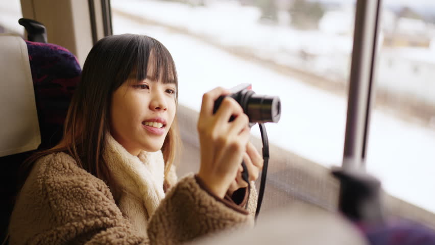 Young Asian woman using digital camera taking picture of beautiful view out of the window during travel on train at sunset. Attractive girl travel Japan on railroad transportation on winter vacation.