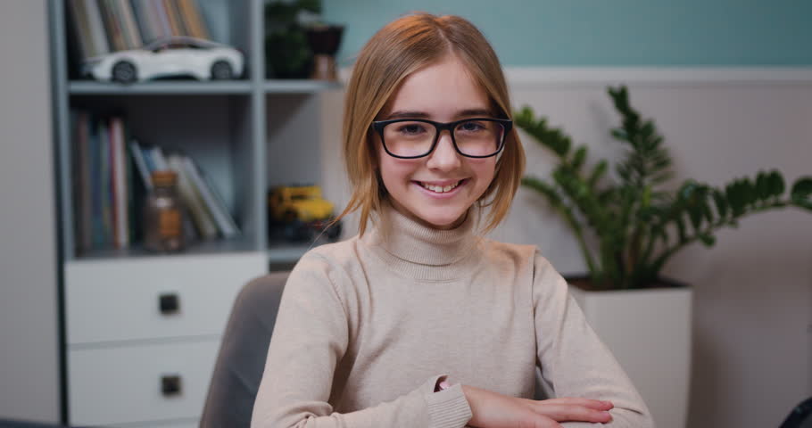 Portrait of smiling caucasian schoolgirl wearing glasses sitting at desk in home class and looking at camera