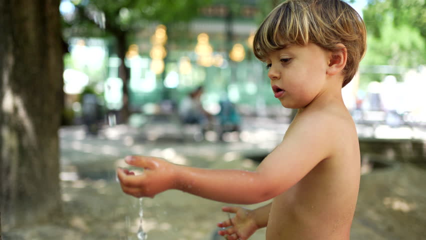 Child washing hands and body at public water park fountain outside in hot summer day. Child heat day refreshment. Lifestyle childhood concept