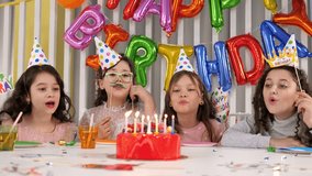 A group of cheerful girls in caps blow out candles together on a cake at a birthday party in a room decorated with balloons. - Powered by Shutterstock - Get 15% off with code: PIKWIZARD15