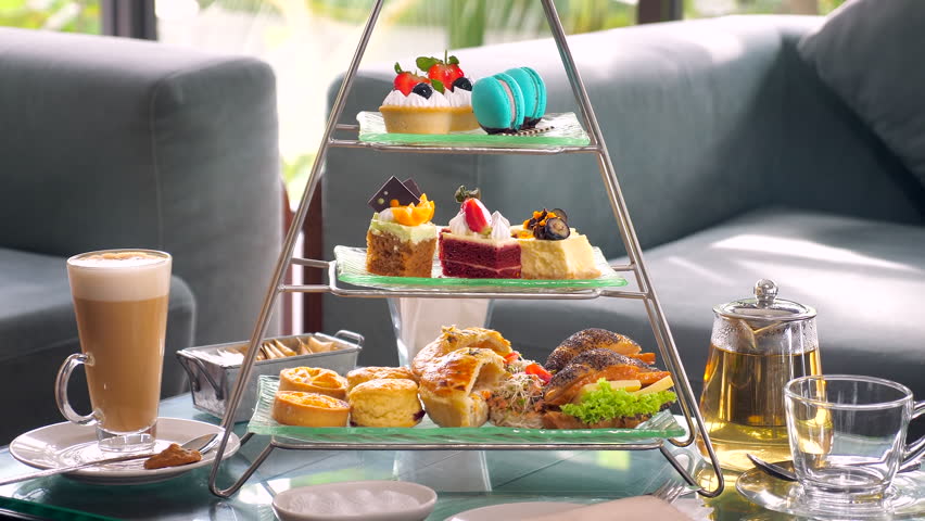 Woman hand take a slice of cake on tiered Afternoon tea display, with cup of coffee and tea on the side, all set against background of tropical restaurant.
