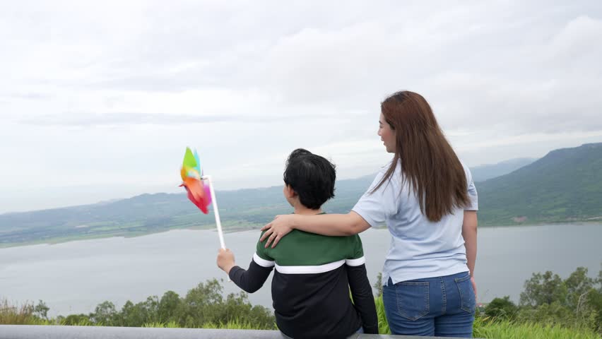 A progressive woman and her son are on vacation, enjoying the natural beauty of a lake at the bottom of a hill while the boy carries a toy windmill.