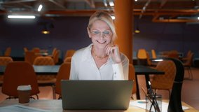 Positive mature businesswoman in eyeglasses holding video conference call with clients, wearing elegant eyewear and classy outfit. Skilled happy saleswoman advertising goods to customers office night