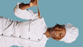 Vertical video: African american cook doing food taste test with spoon, sipping soup for taste testing on camera. Young gourmet chef in apron tasting restaurant meal over blue background, tasty food.