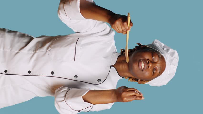 Vertical video: African american cook doing food taste test with spoon, sipping soup for taste testing on camera. Young gourmet chef in apron tasting restaurant meal over blue background, tasty food.
