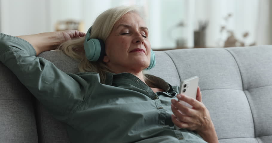 Close up sleepy peaceful mature woman listen music leaned on sofa, holds phone enjoy quality sound through wireless headphones, feels serenity resting on pastime alone at home. Modern tech, relaxation Royalty-Free Stock Footage #1100936525
