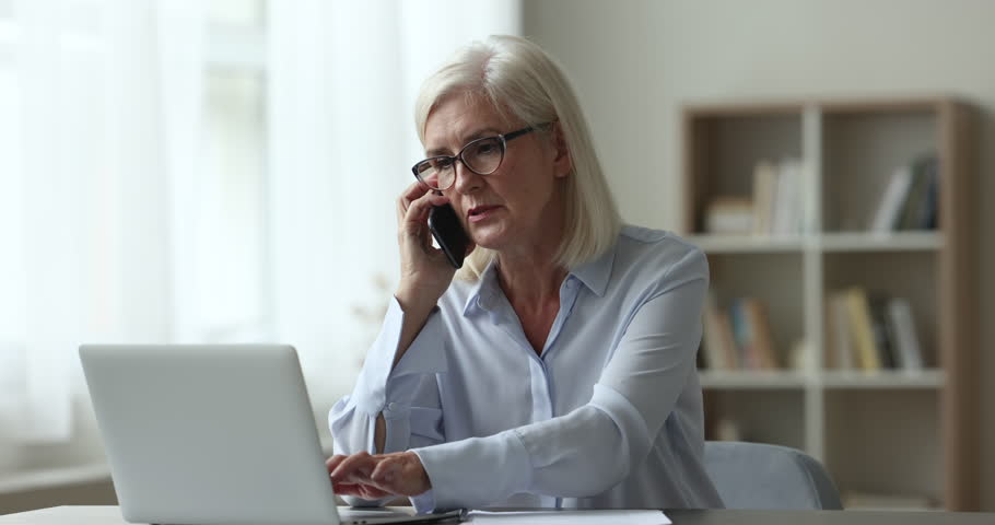 Mature businesslady talking on cellphone seated at workplace desk, share helpful information, solve business, lead formal conversation to client working at homeoffice. Modern wireless tech, phonecall Royalty-Free Stock Footage #1100936535