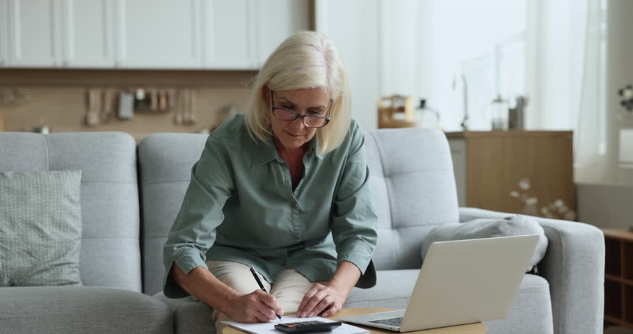 Older woman calculates, writes, pay bills on-line, prepare financial report working from home. Focused mature female, doing accountancy job busy in finances control, manages personal budget use tech Royalty-Free Stock Footage #1100936553