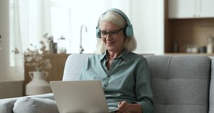 Aged granny sits on sofa with laptop, talk to grandchild use video call app, wear wireless headphones communicates to family living abroad enjoy pleasant warm conversation, involved in virtual meeting