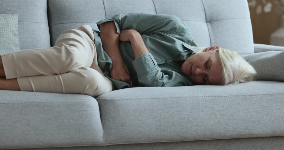 Sick upset aged woman touch belly, suffers from abdomen ache, feels sharp abdominal pain in stomach due to gastritis, pancreatitis symptoms lying frowned on sofa to relieve ache, having health problem | Shutterstock HD Video #1100936609