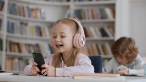 Schoolkids and modern technologies. Cute little girl in headphones web surfing on smartphone at classroom, boy playing video games during break at school, free space