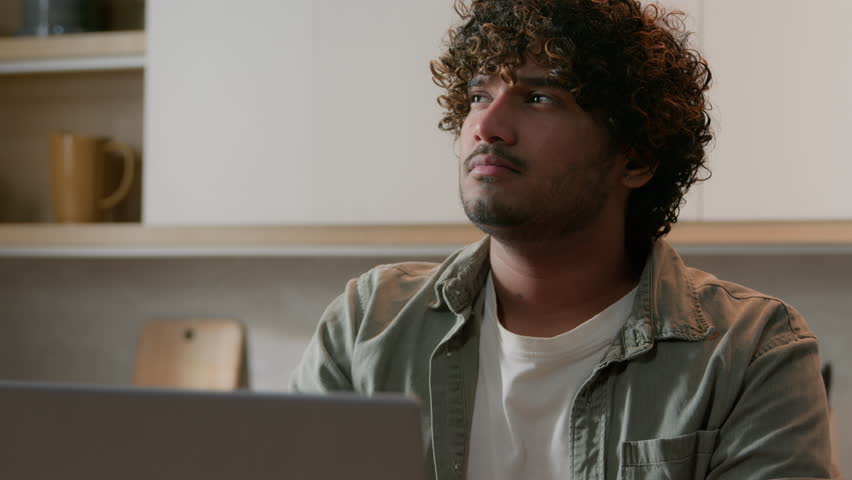 Pensive ethnic guy with curly hair thinking idea typing laptop at kitchen. Thoughtful Indian man search solution working with computer at home. Arabian freelancer think business strategy work online Royalty-Free Stock Footage #1100943599