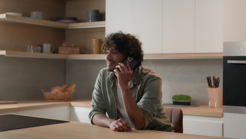 Moving shot laughing smiling cheerful Indian man talking phone in home kitchen. Happy Hispanic guy talk with friends by smartphone Arabian male homeowner speak mobile friendly business conversation Royalty-Free Stock Footage #1100943611