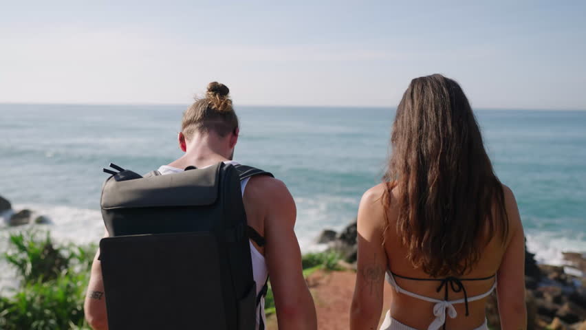 Biracial tourist couple walking towards edge of scenic hill covered with exotic plants above breathtaking view of ocean backview shot. Young adults approach edge of picturescue rock shot from behind