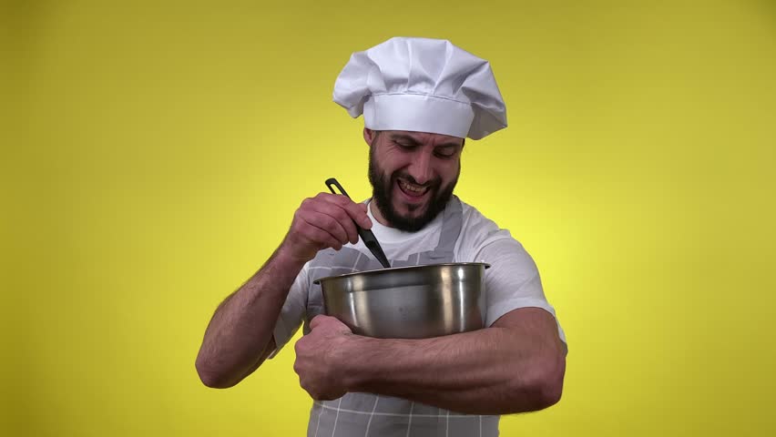 Enthusiastic bearded male cook cooking dessert, whipping egg whites, tasting custard isolated on yellow background. Man chef holding kitchen bowl, beating cream for cake, muffins, cookies indoors.
