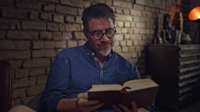 Older man in glasses reading book at home sitting in living room, smiling. 4K video footage. | Shutterstock HD Video #1100966063