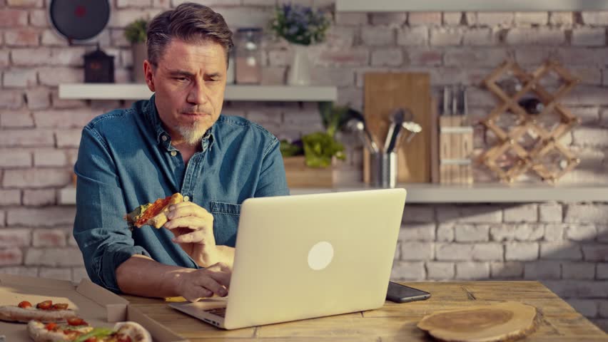 Man working from home on laptop computer, sitting at table in kitchen, eating online ordered pizza. | Shutterstock HD Video #1100966085