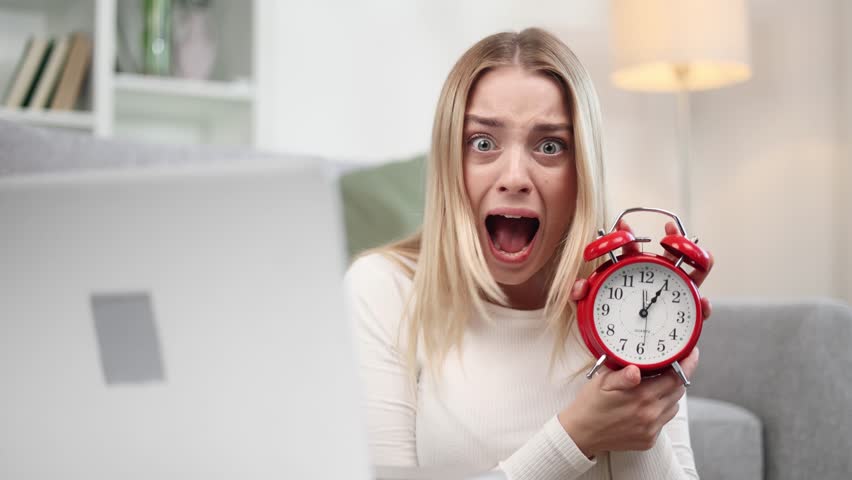 Nervous stressed young blond woman freelancer typing quickly on laptop computer and looking at camera with clock in her hands asking to hurry up scream and warning about deadline at home
