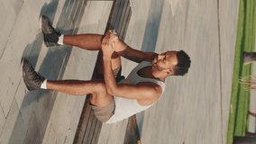 VERTICAL VIDEO: Young bearded male athlete is resting after training, sitting on bench, using smartphone on modern building background