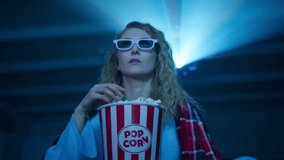 Caucasian woman watching a movie in 3d glasses and eating popcorn at home. Close up of an excited young woman with curly blond hair. High quality 4k footage