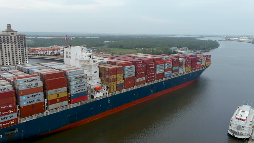 Savannah Georgia USA - 3 1 2023: footage of a large container ship sailing along the rippling green waters of the Savannah River surrounded by hotels, tower cranes and lush green trees on a cloudy day