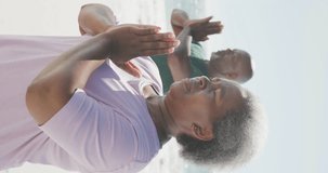 Vertical video of senior african american couple doing yoga at beach, in slow motion. Spending quality time, lifestyle, retirement and holiday concept.