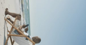 Vertical video of senior african american man using smartphone on deckchair at beach, in slow motion. Spending quality time, lifestyle, retirement and holiday concept.