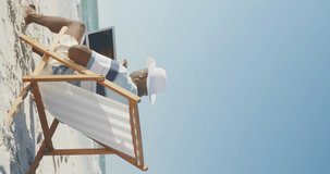 Vertical video of senior african american man using laptop on deckchair at beach, in slow motion. Spending quality time, lifestyle, retirement and holiday concept.