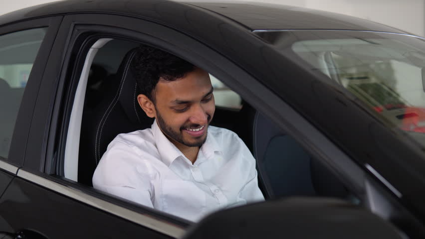 Happy indian man sits in new electric car in shop dealership and celebrate purchase of new vehicle. The man with keys shows emotions of happiness while driving in his new car Royalty-Free Stock Footage #1100977941