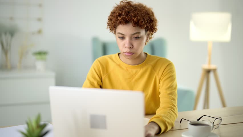 Young woman sits at workplace in home office in front of laptop screen and looks down at desktop with closed eyes and massages bridge of her nose. Tired, sleepy, thoughtful after hard freelance work. | Shutterstock HD Video #1100980629