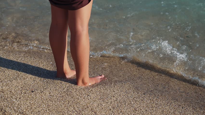 Legs feet of young female walking sand barefoot on the beach. Slim female legs and feet walking along sea water waves on sandy beach. Pretty woman walks at seaside surf. Summer vacation in ocean. | Shutterstock HD Video #1100980689