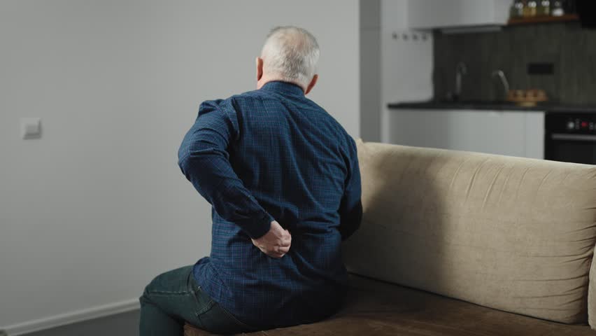 An elderly man sits on the couch at home and has back pain. A man in a shirt at home, suffering from pain | Shutterstock HD Video #1100982265