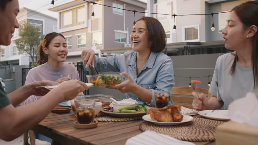 Mom enjoy thai meal cooking for family day home dining at dine table cozy patio. Mum passing serving food to group four asia people young adult man woman friend fun joy relax warm night picnic eating. | Shutterstock HD Video #1100983425