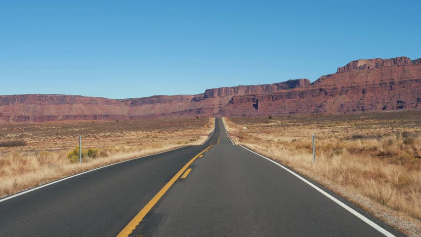 Driving vehicle on empty road going into distance to red mountain rocks in canyon landscape in dried hot desert national park. Highway with black asphalt and orange markings. Slow motion, sunny day