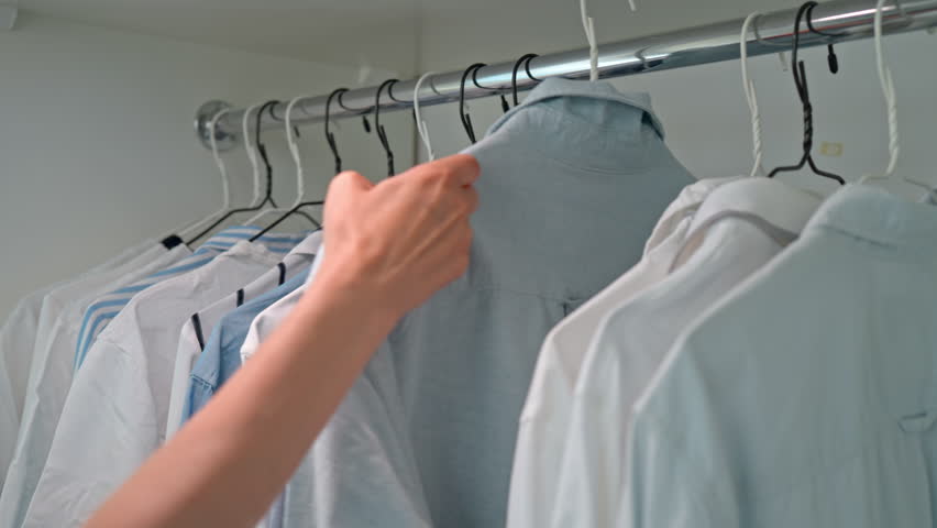 Woman organizes male clothes rack with formal wear blue and white shirts shop assistant holding hanger with casual shirt | Shutterstock HD Video #1100985369