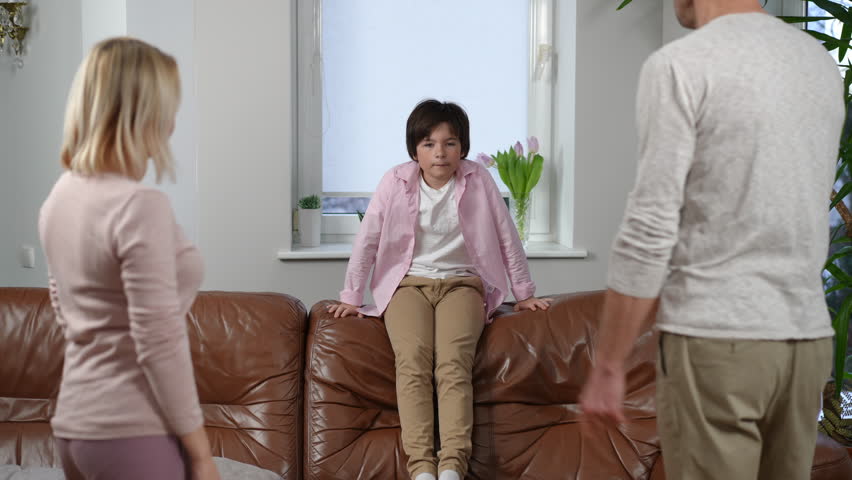 Disobedient boy sitting on couch as unrecognizable parents gesturing scolding kid indoors. Portrait of misbehaving Caucasian son with angry mother and father at home. Individuality and lifestyle Royalty-Free Stock Footage #1100987557