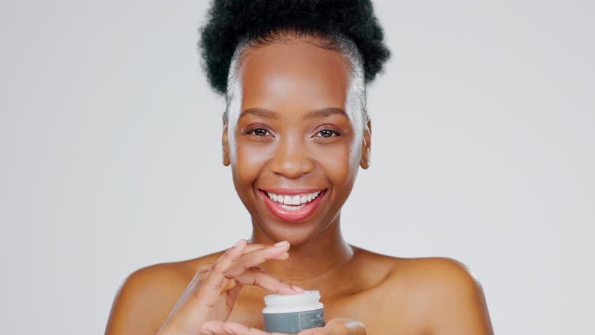 Face, product and skincare with a model black woman in studio on a gray background for beauty treatment. Portrait, wink and lotion with an attractive young female indoor to apply facial moisturizer Royalty-Free Stock Footage #1100990339