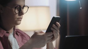 Selective focus of young woman in eyeglasses use cellphone sit at table share text messages in social media, watch internet content take from work on laptop. Tech, e-services, mobile app usage concept