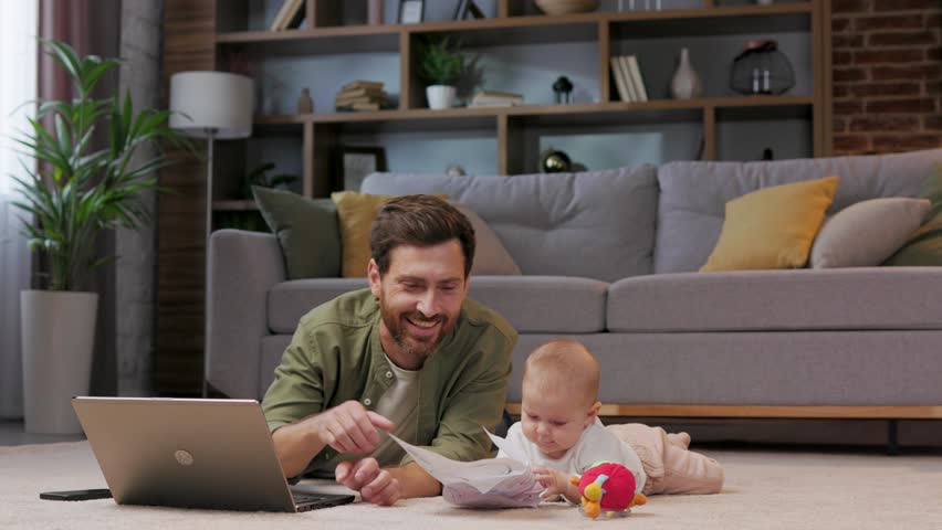 Dad is on maternity leave, he works lying on a mat in an apartment with a laptop and a child. Father care for baby. Father work with child at home. Paternity Leave. Royalty-Free Stock Footage #1100993519