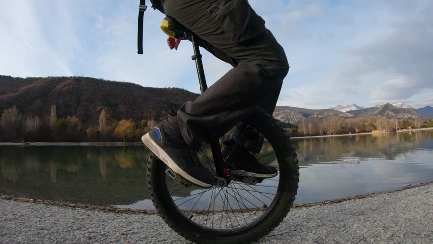 lower part of man body (legs and foot) riding a unicycle on the shore of a lake in Veynes, Hautes Alpes. Lake reflect the background. Side ground view in 60 fps ok for slow motion