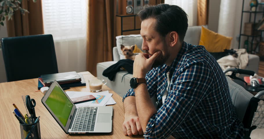 Serious concentrated young man with berd and dark hair sitting in front of coputer thinking solving problem. Man with disabilities in wheelchair working online from distance freelancer concept.
