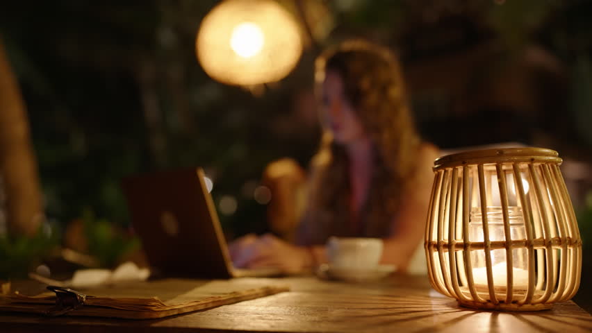 Female freelancer working at night in atmospheric outdoor tropical location closes laptop and leaves the table. Woman sitting at table outside in dark cafe finishes work, takes computer and walks away