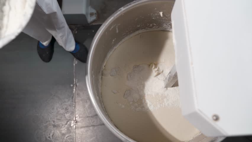 A baker adds flour from a bucket to a dough kneading machine in a bakery production. bakery and industrial food production. | Shutterstock HD Video #1101006291