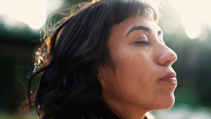 A hispanic latin woman taking a deep breath standing outside in sunlight opening eyes to sky feeling freedom