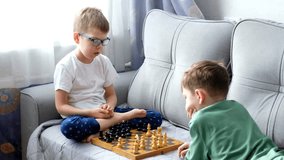 two boys are talking and playing chess sitting on the couch near the window