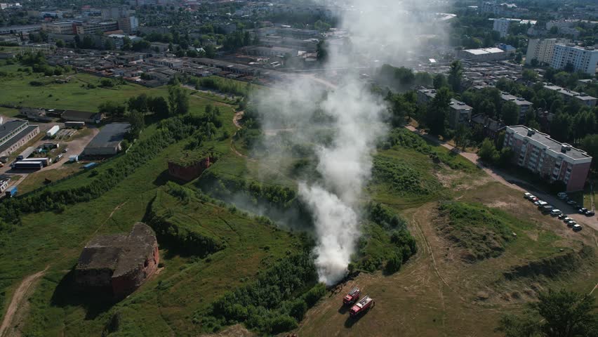 Top view of fire trucks near column of smoke in wasteland next to old fortress across road from residential buildings