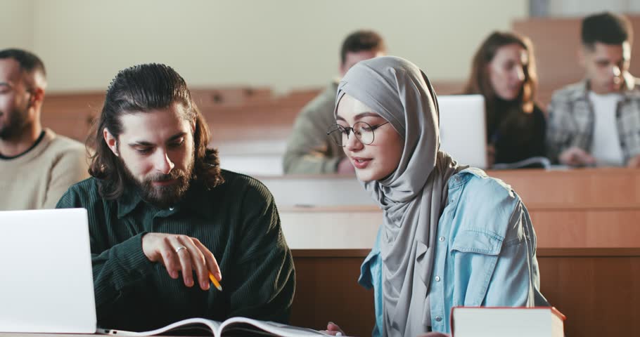Caucasian guy and Arabian girl sitting in auditorium with laptop, talking and working on task at college. Multiethnic male and female at computers discussing studying project at high school. Royalty-Free Stock Footage #1101012807