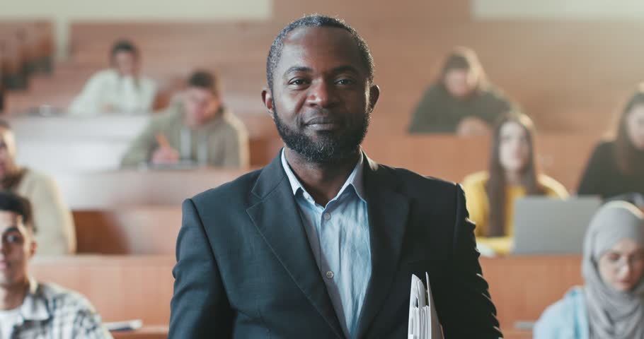 Portrait shot of cheerful African American male professor crossing hands in front of camera and laughing. University. Students on background. Happy man teacher of high school with folder of papers. | Shutterstock HD Video #1101012829